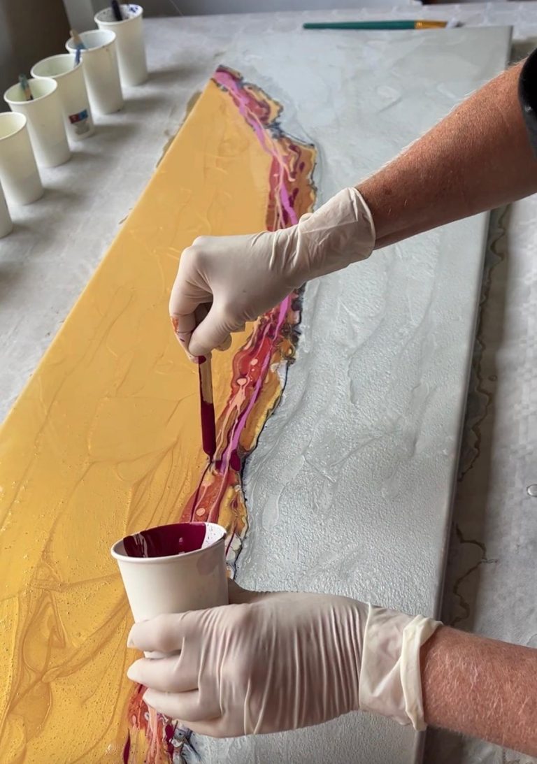 Hand, die violette Farbe auf eine gelbe Fläche aufträgt, mit Bechern im Hintergrund.