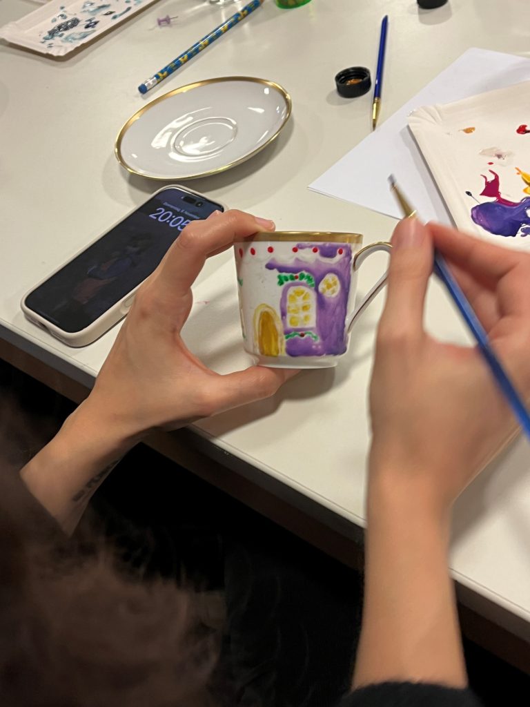 Porzellanmalerei auf Tasse Eine Hand malt ein buntes Motiv auf eine Tasse, mit Pinsel und Farben in der Nähe.