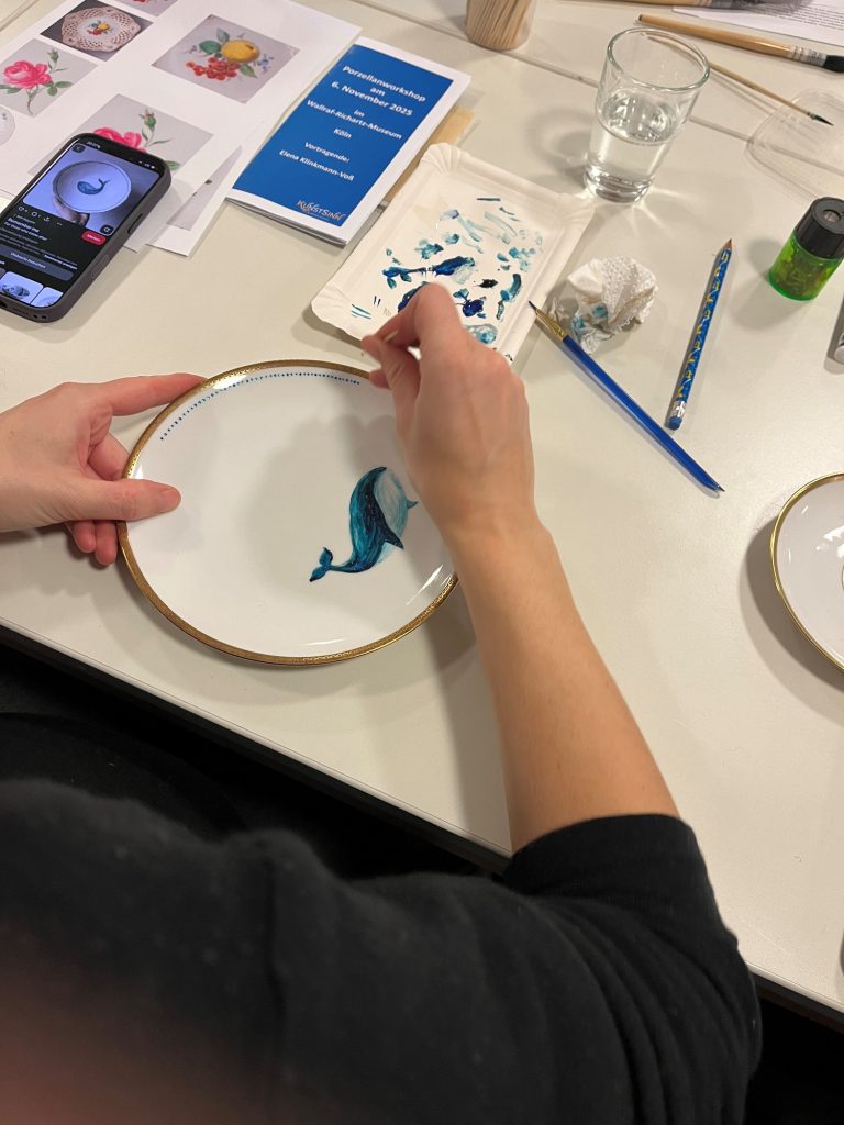 Walfisch auf Porzellanteller Eine Hand malt einen blauen Fisch auf einen weißen Teller, umgeben von Malutensilien.