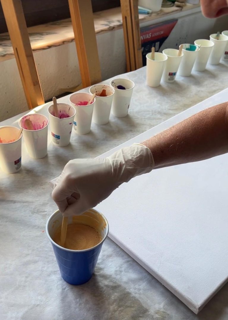 Eine Hand mit Handschuh rührt in einem Becher mit gelber Farbe, dahinter Becher mit Tinte.